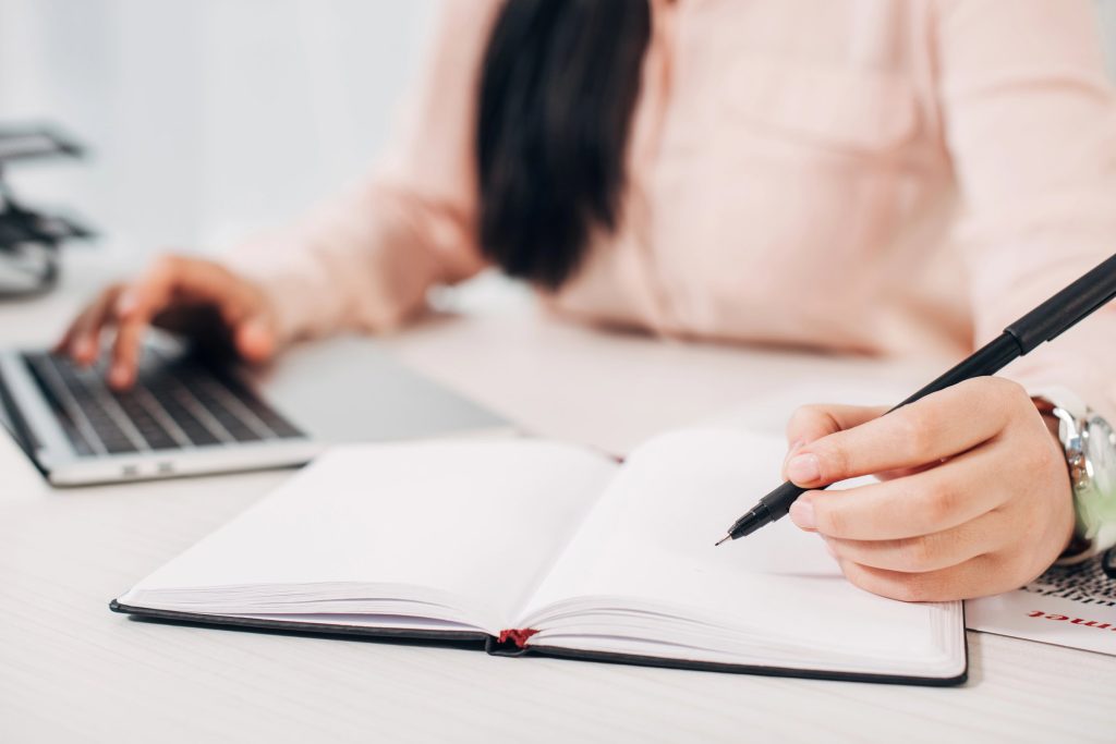 Hand writing a business name and address in a notebook beside a laptop
